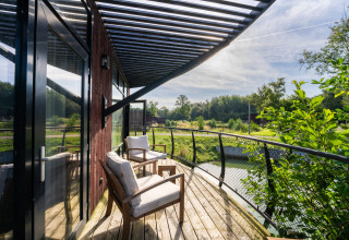 Zonnig terras bij een glampingaccommodatie met comfortabele stoelen en uitzicht op een groene vijver.
