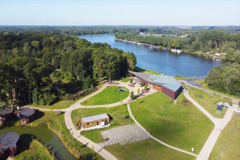 Aerial view of a glamping site by a lake with walking paths, modern lodges, and lush green surroundings.