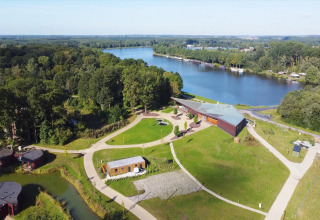 Aerial view of a glamping site by a lake with walking paths, modern lodges, and lush green surroundings.