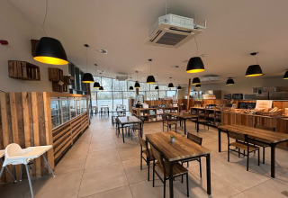 Lyst og moderne caféområde på glampingsted, med træborde, store vinduer og butikshylder med varer.