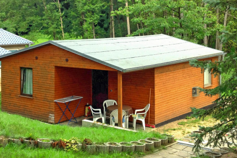 Foto del Bungalow 50 qm 5 Pers., un bungalow di legno con veranda e sedie, immerso nel verde.