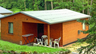 Afbeelding van Bungalow 50 qm 5 Pers., een houten bungalow met terras en stoelen, tussen het groen.