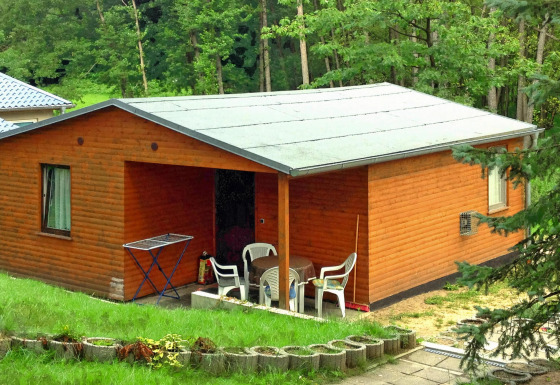 Foto del Bungalow 50 qm 5 Pers., un bungalow di legno con veranda e sedie, immerso nel verde.
