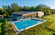 Hébergement glamping avec grande piscine, transats modernes et bâtiment vitré, entouré de verdure et d’arbres.