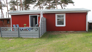 Buitenfoto van een rode lodge genaamd Am Strand met terras, stoelen en grijze omheining op gras.