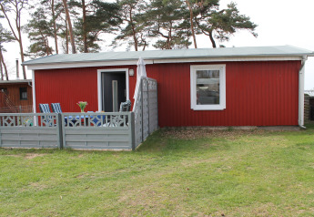 Exterior photo of a red lodge called Am Strand with patio, chairs, and a small fence on green grass.