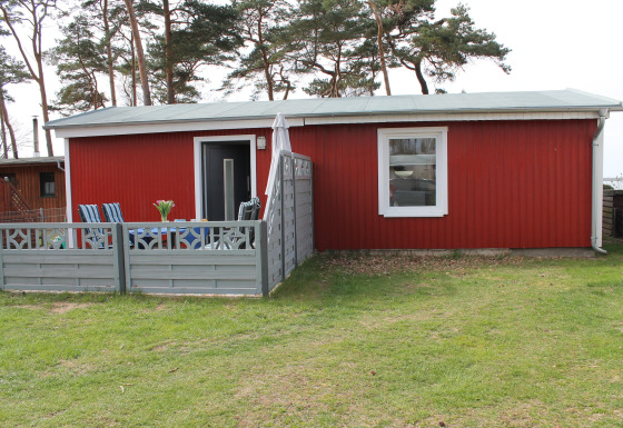 Buitenfoto van een rode lodge genaamd Am Strand met terras, stoelen en hek op een grasveld.