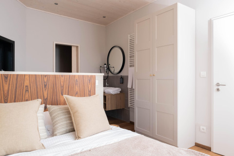 Moderne lodgekamer met bed, houten hoofdbord, lichte kledingkast en ronde spiegel boven de wastafel.