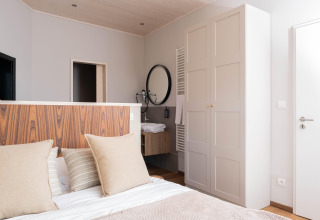 Moderne lodgekamer met bed, houten hoofdbord, lichte kledingkast en ronde spiegel boven de wastafel.