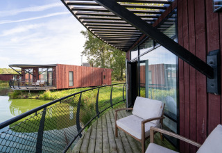Foto van een moderne lodge met houten terras, zitstoelen en uitzicht op water en andere lodges.