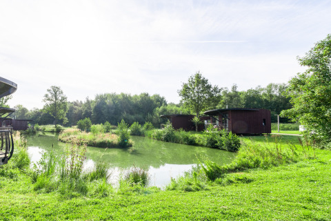 Glampingaccommodatie met moderne huisjes aan een vijver, omgeven door weelderig groen en bomen.