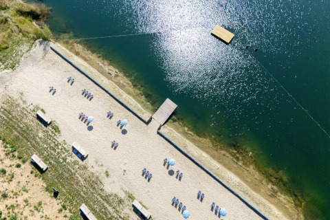 Luftaufnahme eines Sandstrands bei einer Glamping-Unterkunft mit Liegestühlen und Badesteg am See.