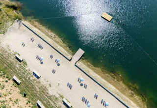 Aerial view of a sandy beach at a glamping site with loungers, umbrellas, and a wooden dock on a lake.