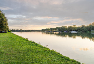 Foto van een rustige plas bij een glamping-accommodatie met groene oever en bomen bij avondlicht.