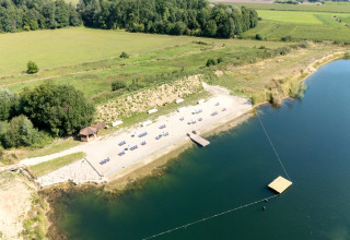 Luchtfoto van een glampinglocatie aan een meer met strandstoelen en omliggende groene velden.