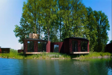 Moderne glampingaccommodatie aan een rustig meer, omgeven door groene bomen en een heldere blauwe lucht.