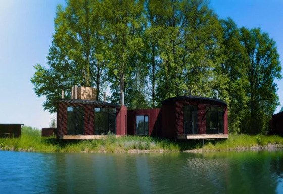 Alojamiento de glamping moderno junto a un lago tranquilo, rodeado de árboles y cielo azul despejado.