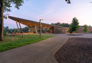 Modern gebouw met houten dak en grote ramen, omgeven door bomen en natuur tijdens zonsondergang.