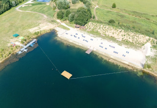 Luchtfoto van een glamping-accommodatie met zandstrand, ligstoelen, meer en vliegtuigen in het groen.