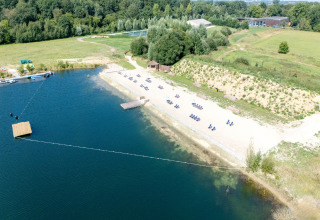 Luchtfoto van een glampinglocatie met zandstrand, ligstoelen, meer en omliggende groene natuur.