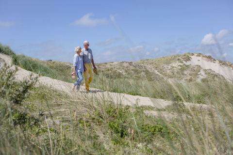 Ouder stel loopt hand in hand door de duinen bij Breeduyn Village 3 in België op een zonnige dag.
