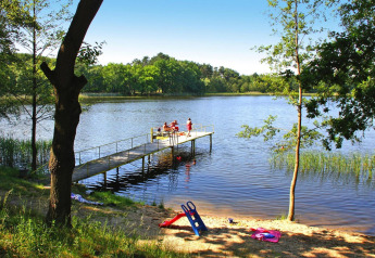 Blick auf einen See mit Steg, Kinder spielen darauf, Spielzeug am Sandstrand, umgeben von Wald.