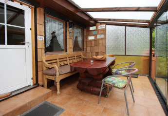 Patio cubierto de madera con paredes de vidrio, mesa rústica, banco, sillas y placas decorativas.