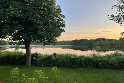 Zicht vanuit een glampingverblijf op een rustige vijver, groen gazon en bomen bij zonsondergang.