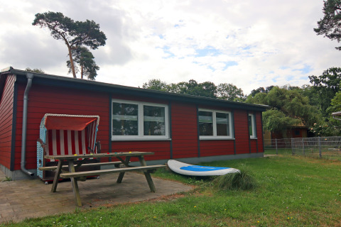 Rood vakantiehuis bij lodge 'Am Strand' in Duitsland, met bank, strandstoel en surfplank buiten.
