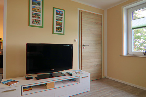 Woonkamer met flatscreen-tv, lichte houten meubels, muurkunst en raam in lodge «Am Strand» in Duitsland.
