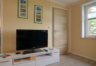 Woonkamer met flatscreen-tv, lichte houten meubels, muurkunst en raam in lodge «Am Strand» in Duitsland.