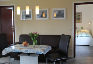 Eetruimte met bruine hoekbank, bloemen op tafel en zicht op slaapkamer in lodge 'Am Strand' in Duitsland.