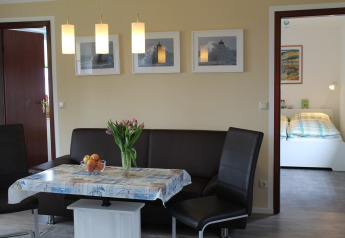 Dining area with brown corner seating, flowers on table and view into bedroom at 'Am Strand' lodge in Germany.
