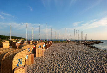 Glamping beach scene with wicker loungers and sailboats docked by a sunny coastline under blue skies.