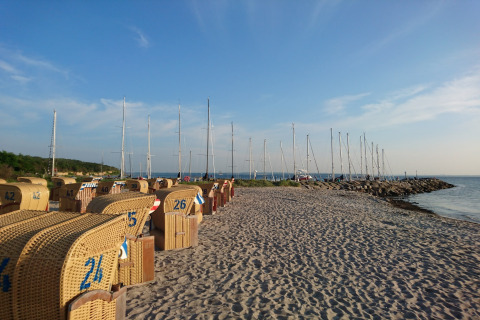 Strand med glamping kurvestole og lystbåde i baggrunden under en klar blå himmel ved kysten.