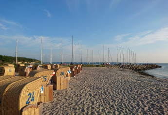 Glamping beach scene with wicker loungers and sailboats docked by a sunny coastline under blue skies.