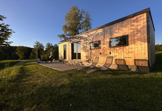 Zonnig zicht op een tiny house met houten gevel en ligstoelen bij SEEBLICK, Eixendorfer See.
