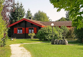 A charming chalet with red shutters, surrounded by lush greenery and trees, under a clear sky.