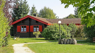 Un bonito chalet con contraventanas rojas, rodeado de césped y árboles, en un entorno verde y soleado.