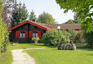 Un bonito chalet con contraventanas rojas, rodeado de césped y árboles, en un entorno verde y soleado.