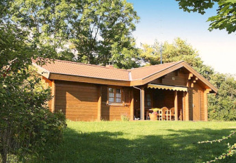 Wooden chalet surrounded by trees and greenery, with a covered porch and outdoor seating area visible.