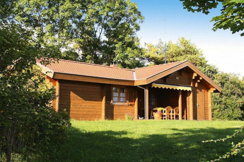 Chalet in legno immerso nel verde, circondato da alberi, con veranda coperta e tavolo esterno.