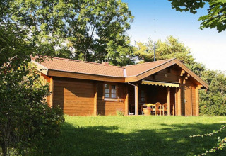 Wooden chalet surrounded by trees and greenery, with a covered porch and outdoor seating area visible.