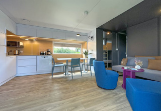 Moderne tiny house met keuken, eettafel, blauwe stoelen, bank en lichte houten vloer.