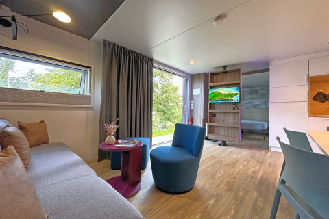 Modern tiny house living room with sofa, blue chair, small table, TV, large window, and bedroom access.