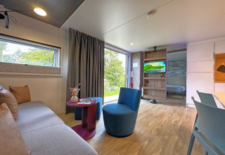 Sala de estar en tiny house moderna con sofá, silla azul, mesa pequeña, televisor, ventana grande y dormitorio.