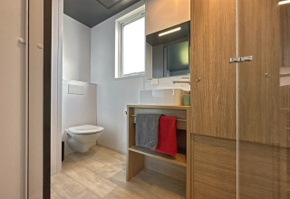 Modern tiny house bathroom with a wall-mounted toilet, wood cabinetry, towels, and a large window.