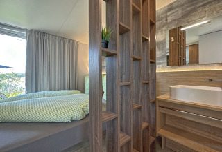 Tiny house met modern interieur, slaapkamer en badkamer gescheiden door houten rek, veel daglicht en stijl.