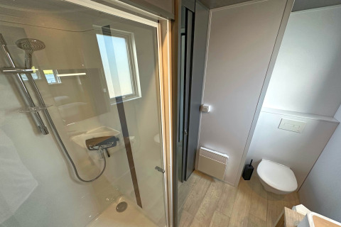 Bagno moderno in una tiny house con doccia in vetro, WC sospeso, finestra e pavimento in legno.