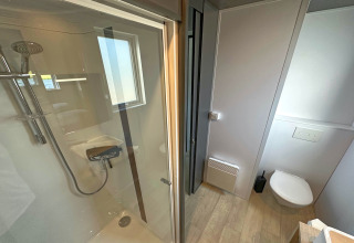 Baño moderno en una tiny house con ducha de cristal, inodoro suspendido, ventana y suelo de madera.